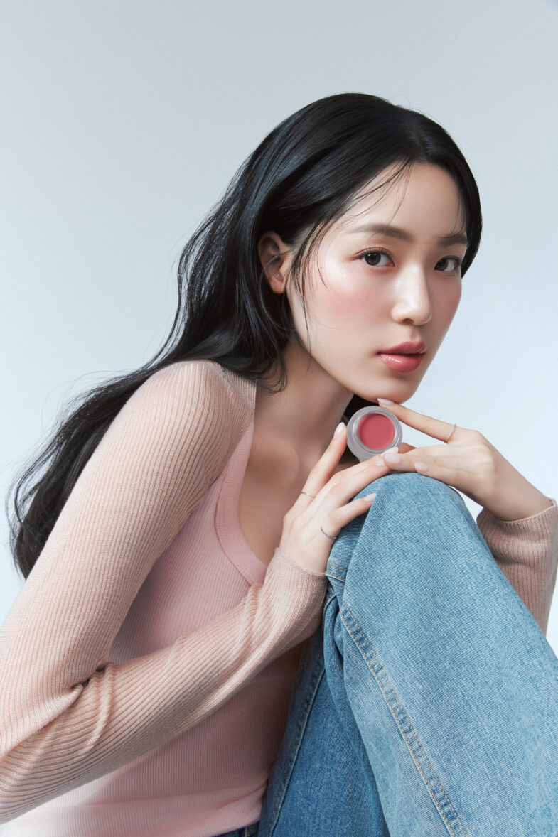 A person with long dark hair holds a pink makeup product, wearing a pink top and jeans against a light background.