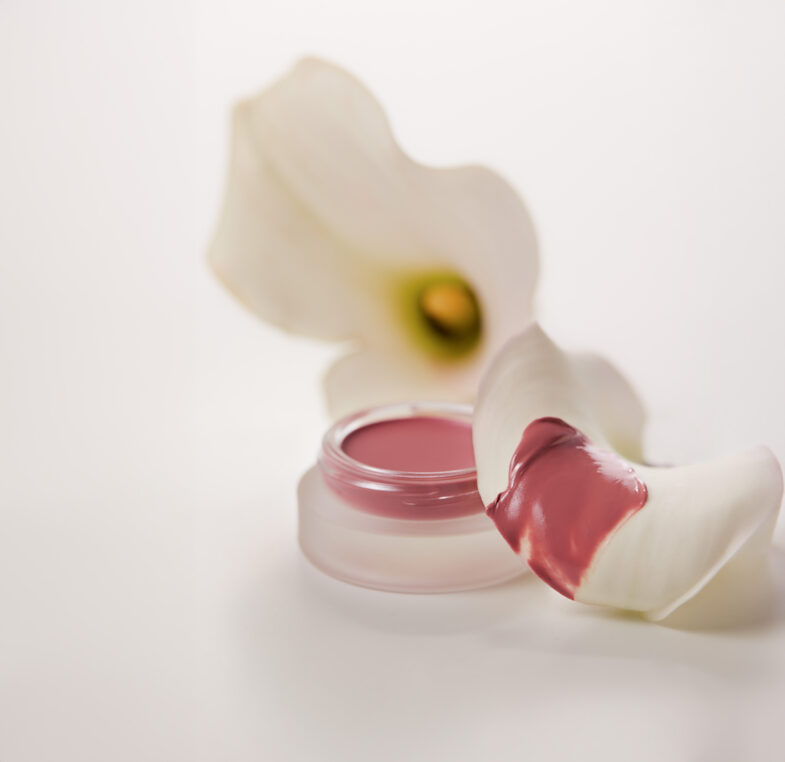 A small container of pink lip balm next to a white flower petal stained with pink, against a soft background.