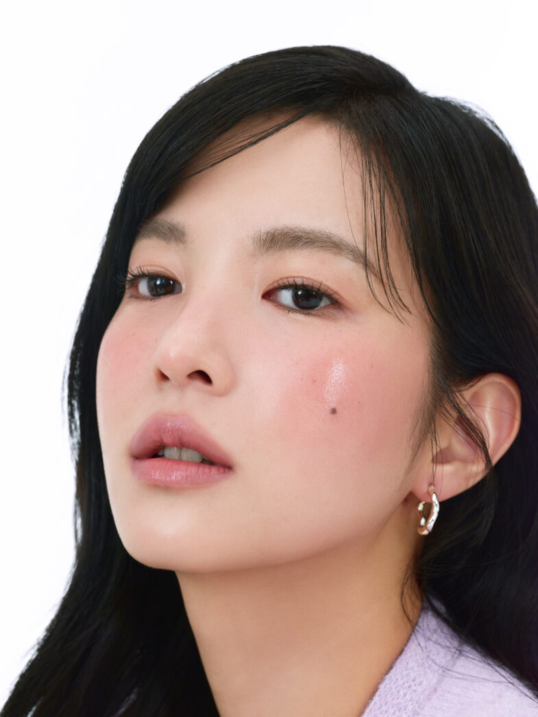 Person with long black hair and hoop earrings against a white background, displaying natural makeup and a beauty mark.