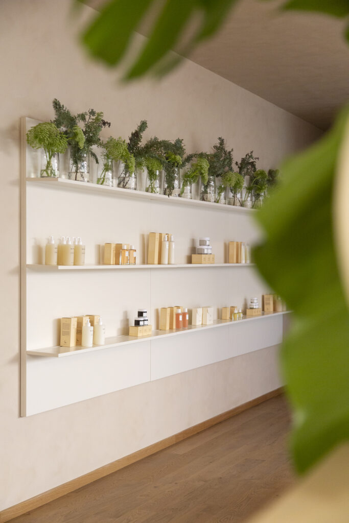 Police s kozmetičkim proizvodima na kremastim zidovima, ukrašene staklenim vazama s zelenilom na gornjoj polici, s drvenim podom u primjeni.