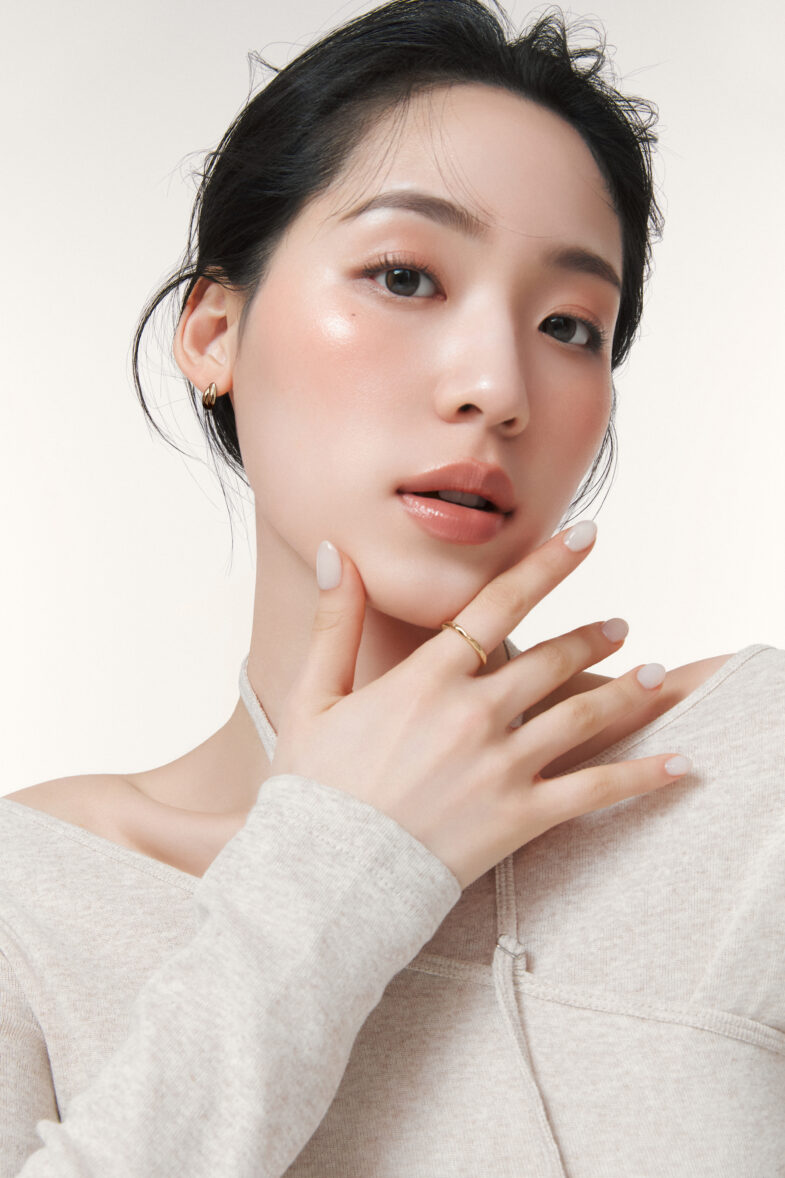 Person with glowing makeup and gold hoop earrings, wearing a light beige top, with a thoughtful pose.