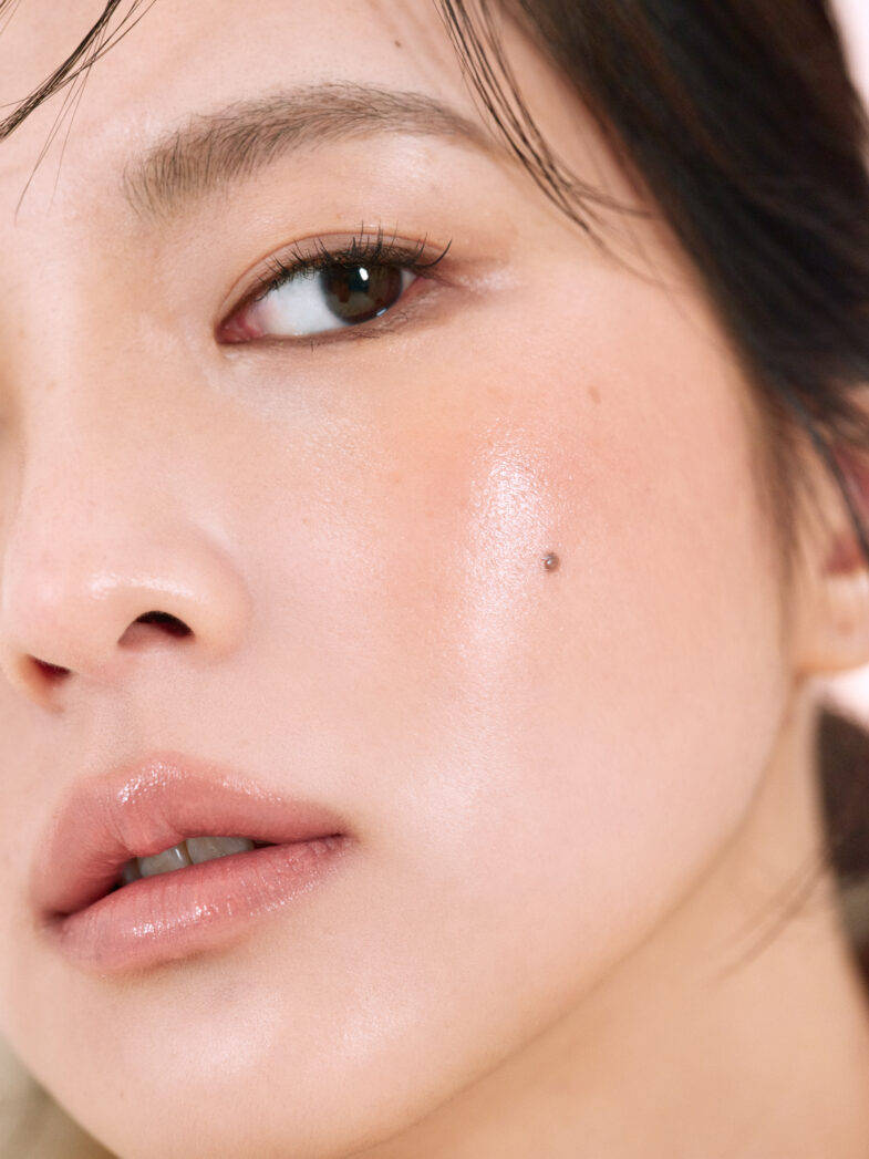 Close-up of a woman's face showcasing smooth skin, glossy lips, and a beauty mark on her cheek.