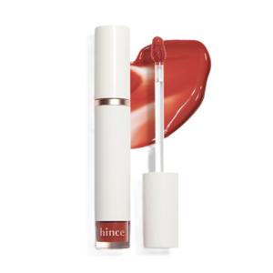 A tube of hince lipstick with a white cap, accompanied by an applicator and a smear of reddish-orange color.